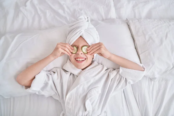 Havlu Türbanlı Bornozlu Gülümseyen Çocuğun Gözlere Salatalık Dilimleri Sürerken Spa — Stok fotoğraf
