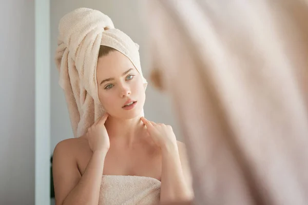 Young calm woman with towel on head admiring natural beauty while standing in bathroom and looking in mirror after taking shower