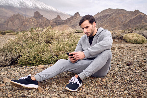 Side view of bearded concentrated male in activewear texting message on mobile phone while resting on stony ground near volcano Teide in Tenerife in Canary Islands in Spain
