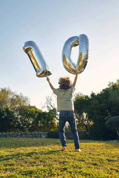 太陽の光の下で芝生の上で誕生日を祝う腕や装飾的な風船を上げた無名の子供のバックビュー — ストック写真