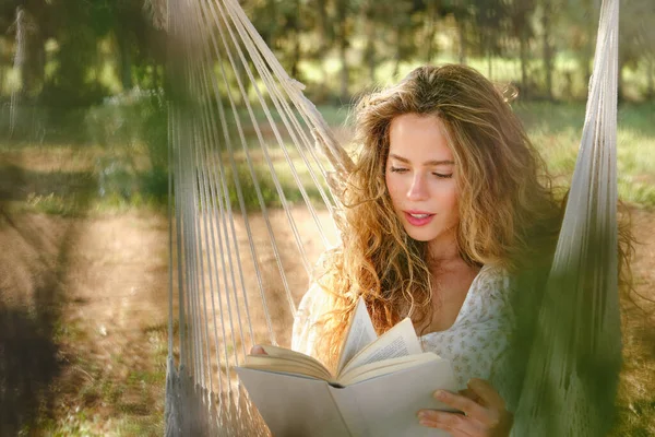 Młoda Kobieta Kręconymi Włosami Siedząca Hamaku Czytająca Ciekawą Książkę Słoneczny — Zdjęcie stockowe