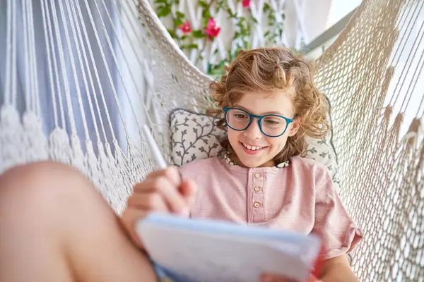 Uśmiechnięte Dziecko Okularach Odpoczywające Hamaku Dzianiny Poduszką Piszące Pamiętniku Ołówkiem — Zdjęcie stockowe