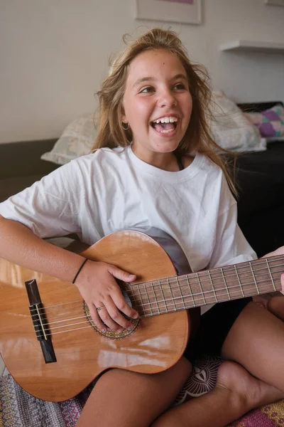 リビングルームの床に座っている間 歯ぎしりの微笑みと白いTシャツでアコースティックギターを演奏する興奮した肯定的な少女 — ストック写真