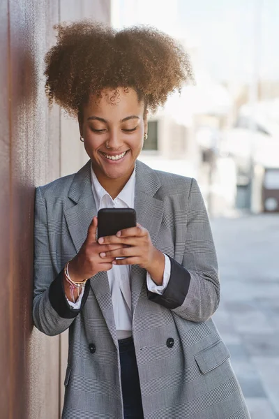 Positive African American female entrepreneur in elegant jacket texting on mobile phone against blurred background of street