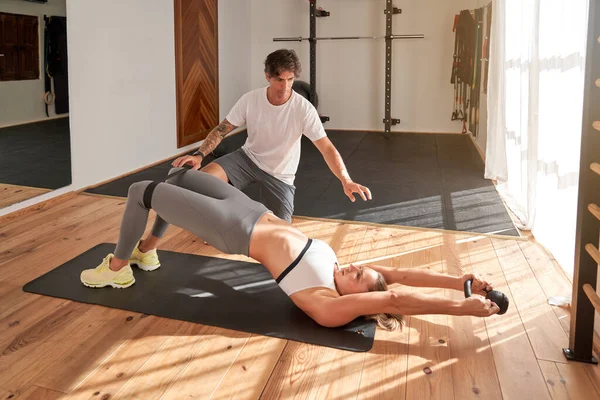 Full body of male instructor helping young female athlete doing exercises with resistance band and kettlebell during workout in modern fitness club