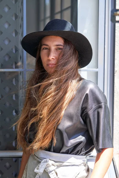 身穿时髦服装 头戴帽子 自信的年轻女性站在窗前看着相机 — 图库照片
