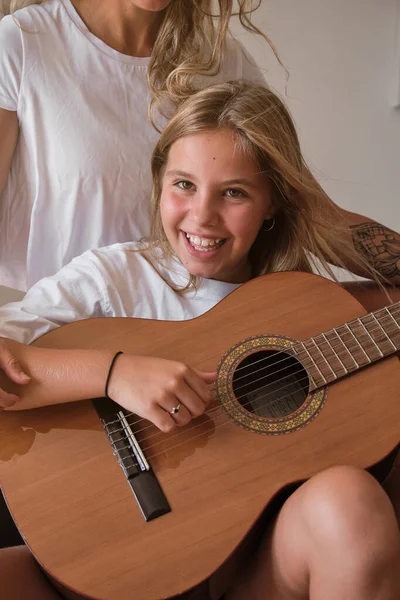 ブロンドの女の子がギターを弾きながら 部屋のソファーに座っている女性とカメラで微笑んでいる姿の垂直写真 — ストック写真
