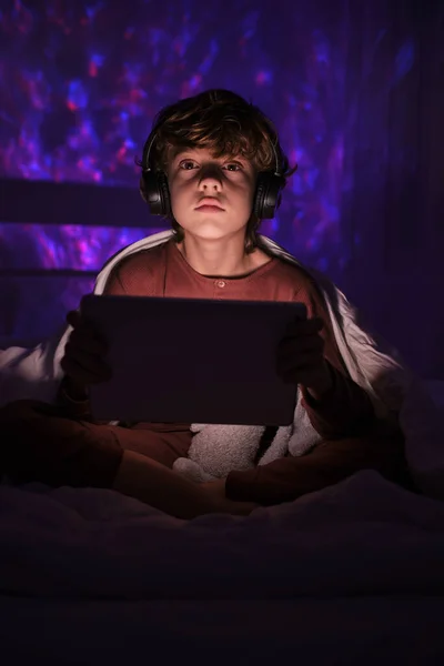 헤드폰을 바라보고 귀여운 밤중에 아파트에서 불빛을 받으며 침대에 현대식 태블릿을 — 스톡 사진