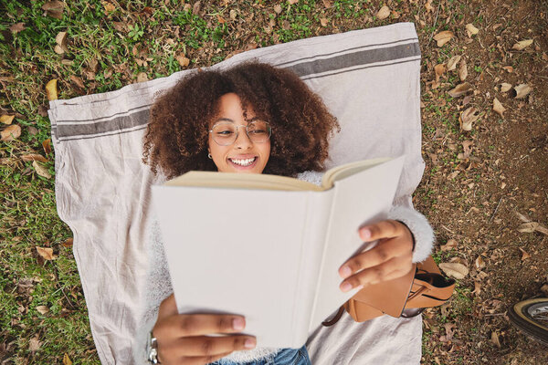 Вид сверху чернокожей женщины с афро-прической, читающей интересную историю и улыбающейся, отдыхая на одеяле на траве в природе
