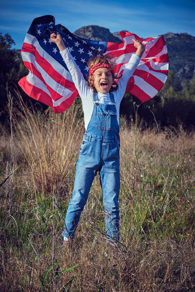 Smiling preteen boy in jeans overall standing in grassy meadow with raised arms with flag of United States of America