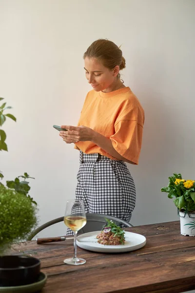 家で居心地の良いキッチンでワインとステーキタータルと木製のテーブルの近くに立っている間 電話でテキストメッセージカジュアル服の正の若い女性 — ストック写真