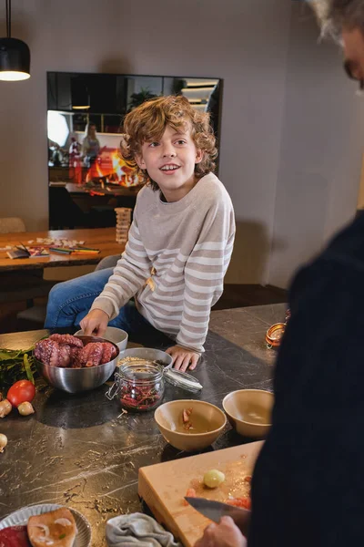 Curly Uśmiechnięty Chłopiec Swetrze Siedzi Szerokiej Blacie Kuchennej Pełnej Naczyń — Zdjęcie stockowe