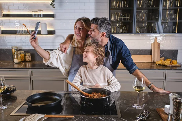 Cheerful diverse parents in aprons cooking together with preteen son in kitchen and taking self portrait on smartphone