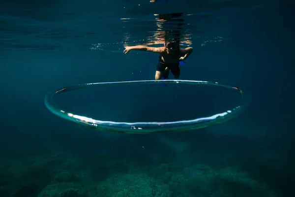 サンゴ礁近くのトロイダルバブル近くの海水の下に浮かぶショートパンツとシュノーケリングマスクの若いシャツレス男性ダイバーのフルボディ — ストック写真