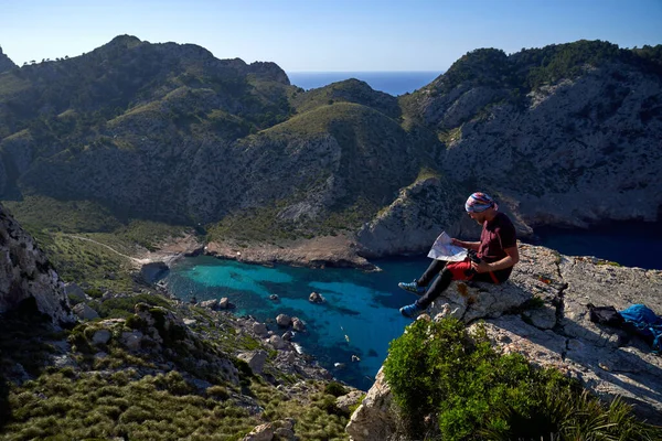 在阳光灿烂的日子里 身穿休闲装的男性游客坐在岩石悬崖上 阅读纸质地图的侧视图 — 图库照片