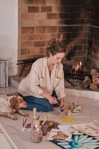 专注的女艺术家坐在靠近宠物的地板上 用专业的画笔在面料上画画 并在创造性的工作空间中绘画 — 图库照片