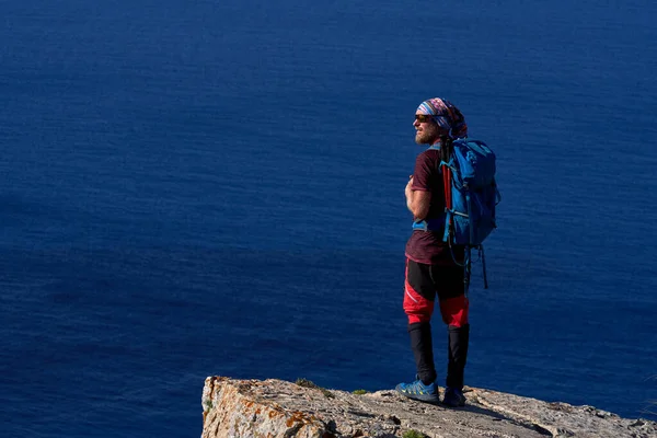 バックパック付きの男性ハイカーの完全な長さのバックパック岩の崖の上に立って離れて海に対して見て — ストック写真