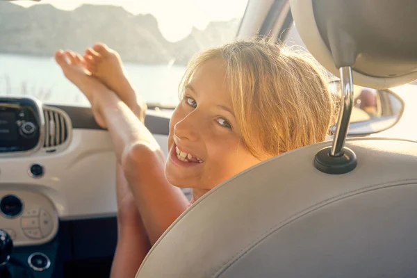 車の前の彼女の足で車の中に座っているブロンドの女の子と背景に海とカメラに直面することになります — ストック写真