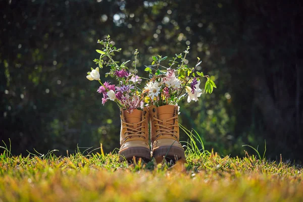 夏の日に背景がぼやけて緑豊かな木々と日当たりの良い芝生の芝生の上でカラフルな野花と茶色のブーツのペア — ストック写真