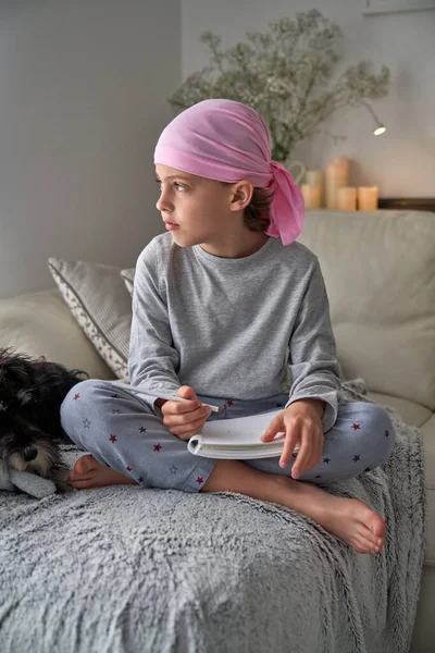 フルボディのPajamaとピンクのスカーフシンボルの乳がん意識座っクロス足と離れて見ている間にノートブックで書く近くミニチュアSchunazer嘘上のソファ — ストック写真