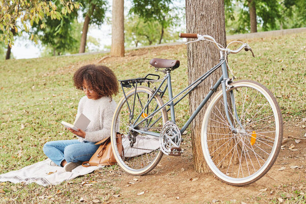 Все тело сконцентрированной афроамериканки в повседневной одежде сидит на одеяле возле велосипеда и читает интересную историю в зеленом парке