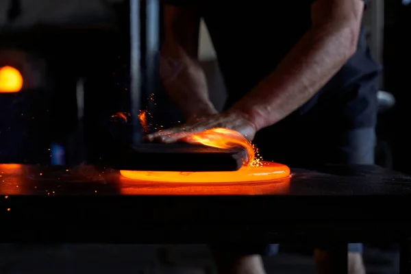 工場のテーブルで熱いオレンジ色のガラスを切る黒いカジュアルな服の熟練した認識できない男性労働者の手 — ストック写真