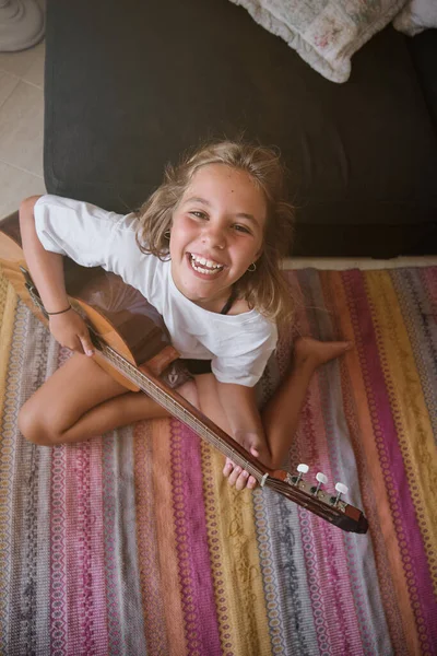 カーペットの上に座ってアコースティックギターを演奏しながら カメラを見ながら笑顔でカジュアルな服を着た幸せな少女の上から — ストック写真