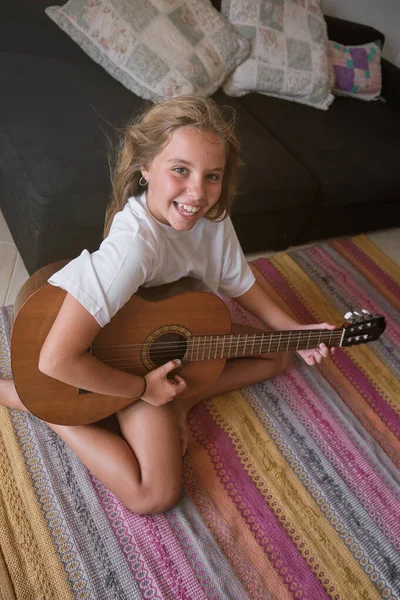 優秀な女の子の上から笑顔で 家のリビングルームでアコースティックギターで曲を演奏しながらカメラを見る — ストック写真