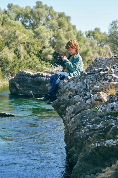 自然の岩の海岸の端に座っている間に川の写真を撮る陽気な少年の完全なボディサイドビュー — ストック写真