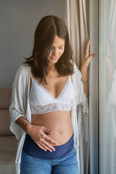自宅の窓の近くに立って 妊娠中の腹に触れながら 白い下着とジーンズの目を閉じる平和な女性 — ストック写真
