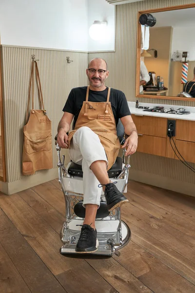Cuerpo Completo Barbero Barbudo Delantal Sentado Silla Mirando Cámara Mientras — Foto de Stock