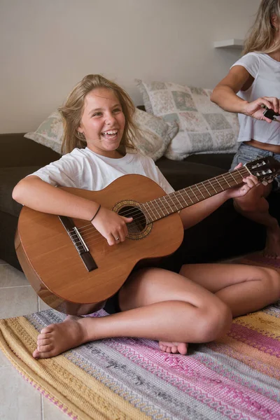 リビングルームのソファーの近くのカーペットに座って 週末にアコースティックギターを演奏しながら カジュアルな服で笑う少女の全長 — ストック写真