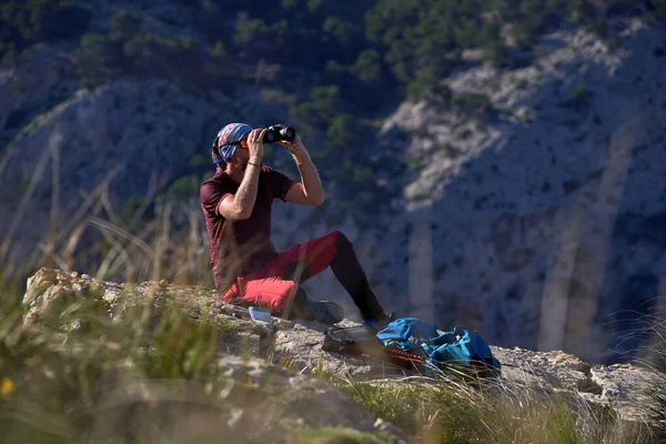 サイドビューフルボディの男性ハイカーは岩の崖の上に座って 晴れた日に山に対して双眼鏡を通して離れて見て — ストック写真