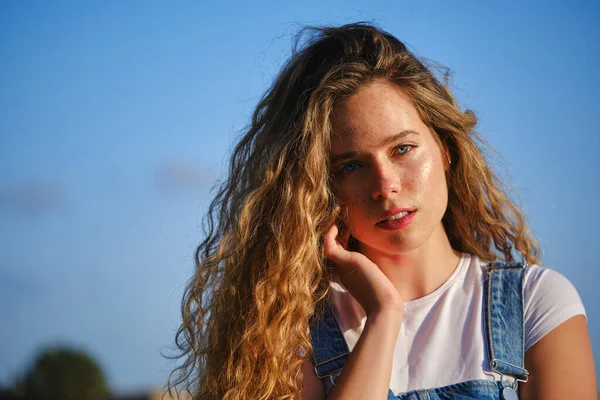 夏の田舎の青空の下でカメラを見ながら顔を触っているカジュアルな服を着た美しい若い女性 — ストック写真