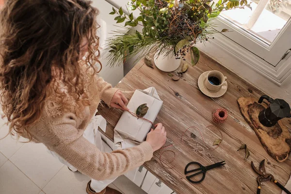 从上到下站在木制桌旁 拿着咖啡和花束 装饰着用叶子包裹的礼物 准备在家里庆祝节日 那是一个难以辨认的有创意的女人 — 图库照片