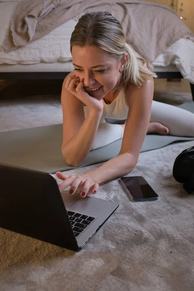 Smiling female athlete in sportswear sitting on mat and searching for online tutorial while preparing for yoga class in bedroom at home