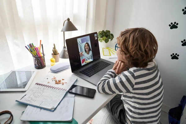 テーブルに座り 部屋で宿題をしている間 ネットブックを介して友人とビデオチャットをするカーリーヘアを持つ19歳の少年を横に見る — ストック写真