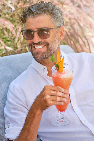 Happy smiling bearded male traveler wearing sunglasses holding cocktail glass in hand while sitting on mattress of wooden bench and looking away