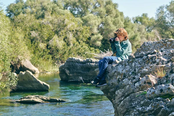 聚精会神的男孩坐在绿树成荫的近岸石质海岸边 用照相相机拍照 全景全景 — 图库照片