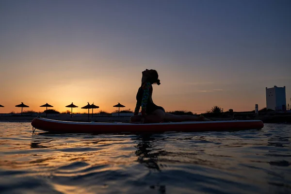 日落时分 身穿泳衣的年轻女子在海滨的船舷上做瑜伽的同时 还摆出了半鸽的姿势 — 图库照片
