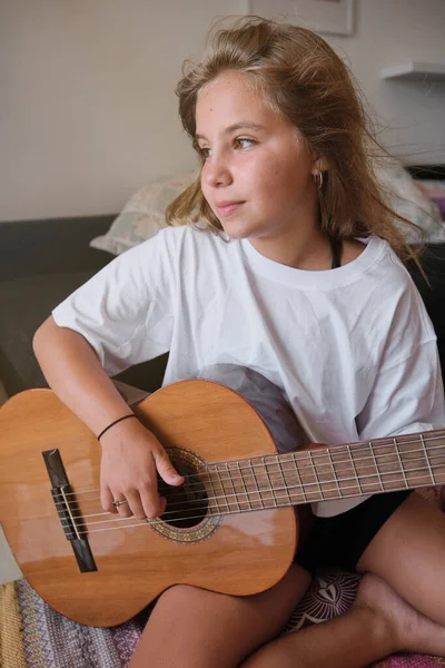 ソファー付きの家の部屋でギターを演奏しながら 気を散らした表情を持つブロンドの女の子の垂直写真 — ストック写真