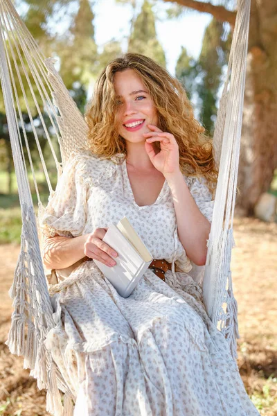 明るい夏の日にハンモックでリラックスしながら本の感動的な顔と笑顔でカメラを見て陽気な若い女性 — ストック写真
