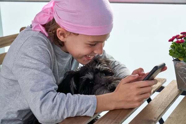 Side view of kid in pink headband sitting at wooden table with small dog and browsing mobile phone with smile