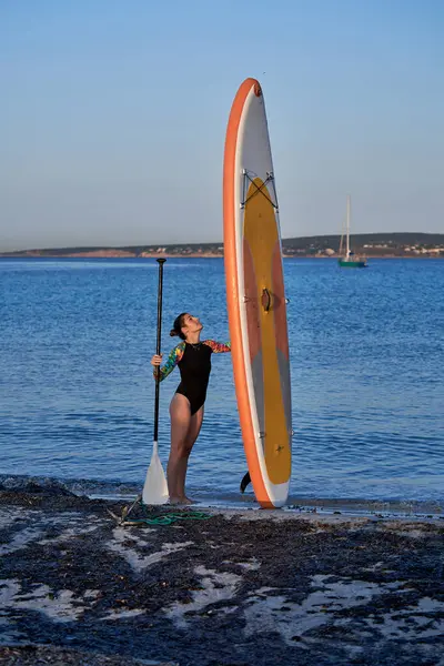Side view full body of female surfer in swimwear carrying paddle and standing with vertical SUP board on seashore against picturesque scenery