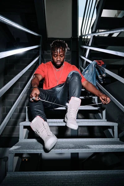 Full body of young African American male in trendy outfit sitting on stairs and tying shoelaces on fashionable sneakers