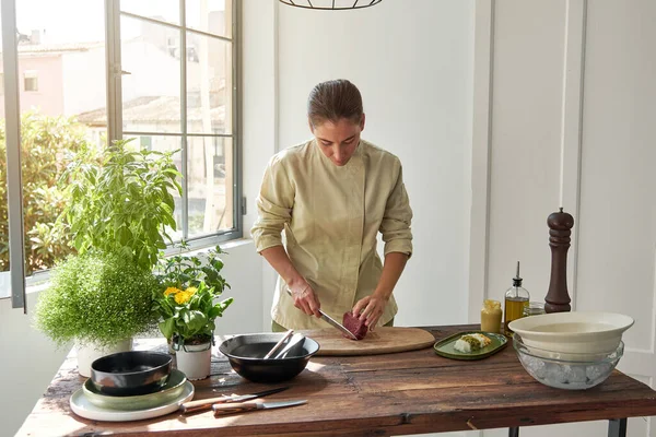 家庭の台所で昼食を調理しながら 木製のテーブルに立って まな板の上に牛肉を切断する統一されたプロの女性シェフ — ストック写真