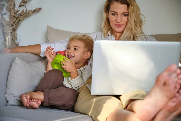 大人の母親は ラップトップでビデオを見て 自宅のリビングルームでソファーで休んでいる間 緑のボウルで笑顔の息子を抱きしめます — ストック写真