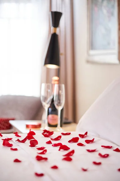 Romantic atmosphere interior of hotel suite room with bed covered with white bedclothes and decorated with scattered red rose petals and bottle of champagne with glasses placed on bedside table