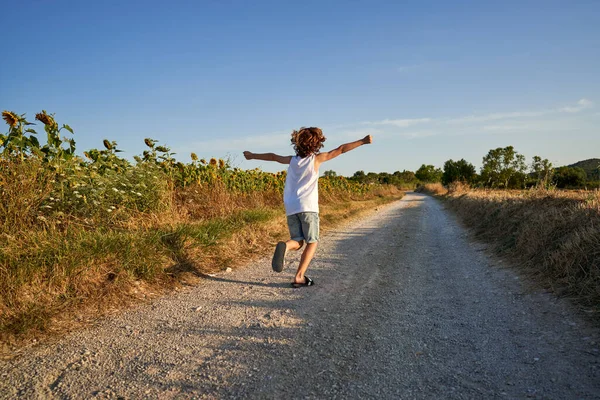 夏の日にひまわり畑近くの田園道を走っている間に腕を広げて飛ぶ巻き毛のある匿名の子供の背中の景色 — ストック写真