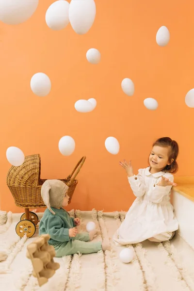 オレンジ色の壁とイースターエッグで部屋に座っているドレスで愛らしい赤ちゃんの少女と幼い姉妹のフルボディ — ストック写真